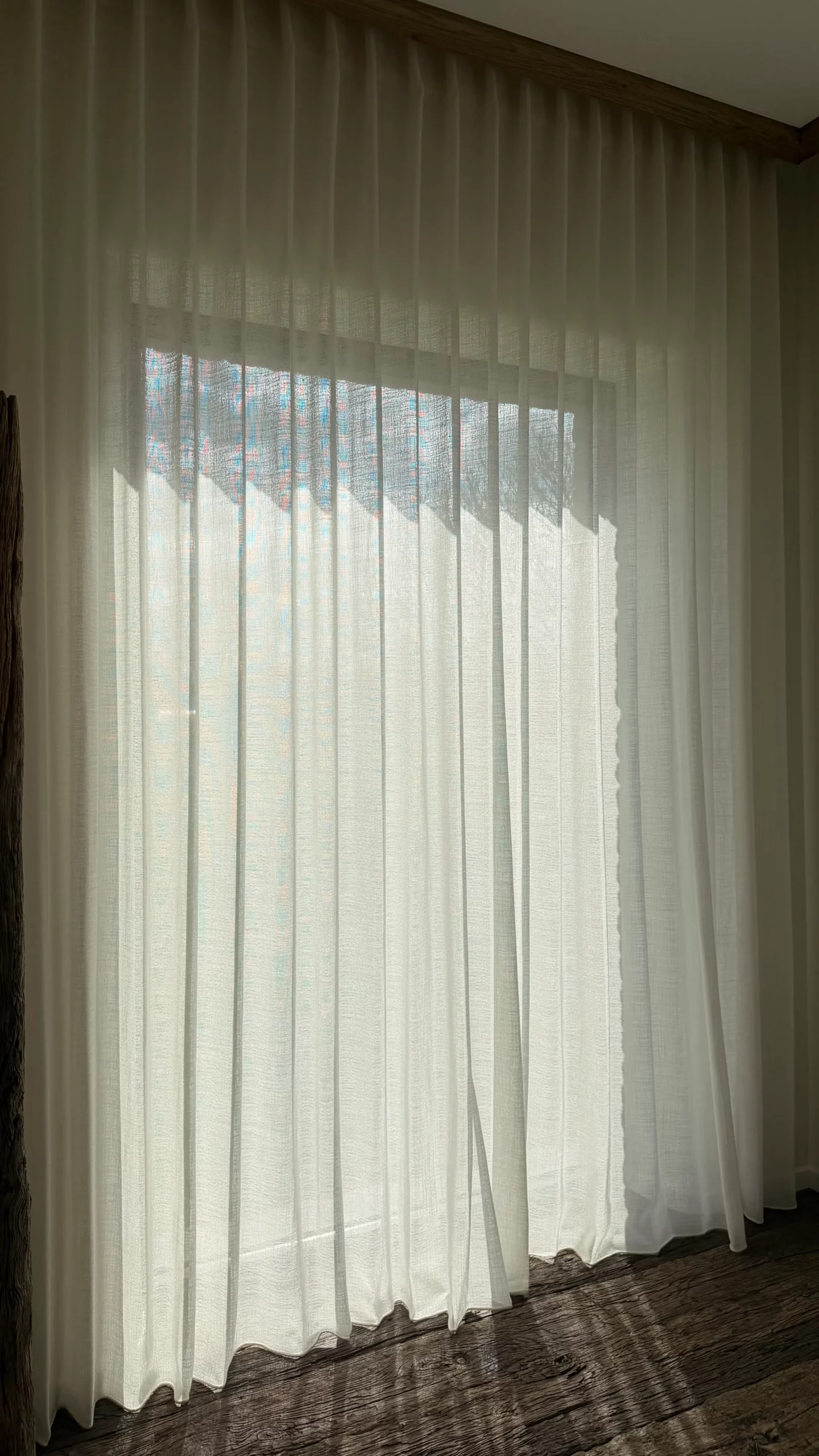 Wit vitragegordijn met golfplooi voor een raam, met zonlicht dat door de stof valt in een kamer met houten vloer