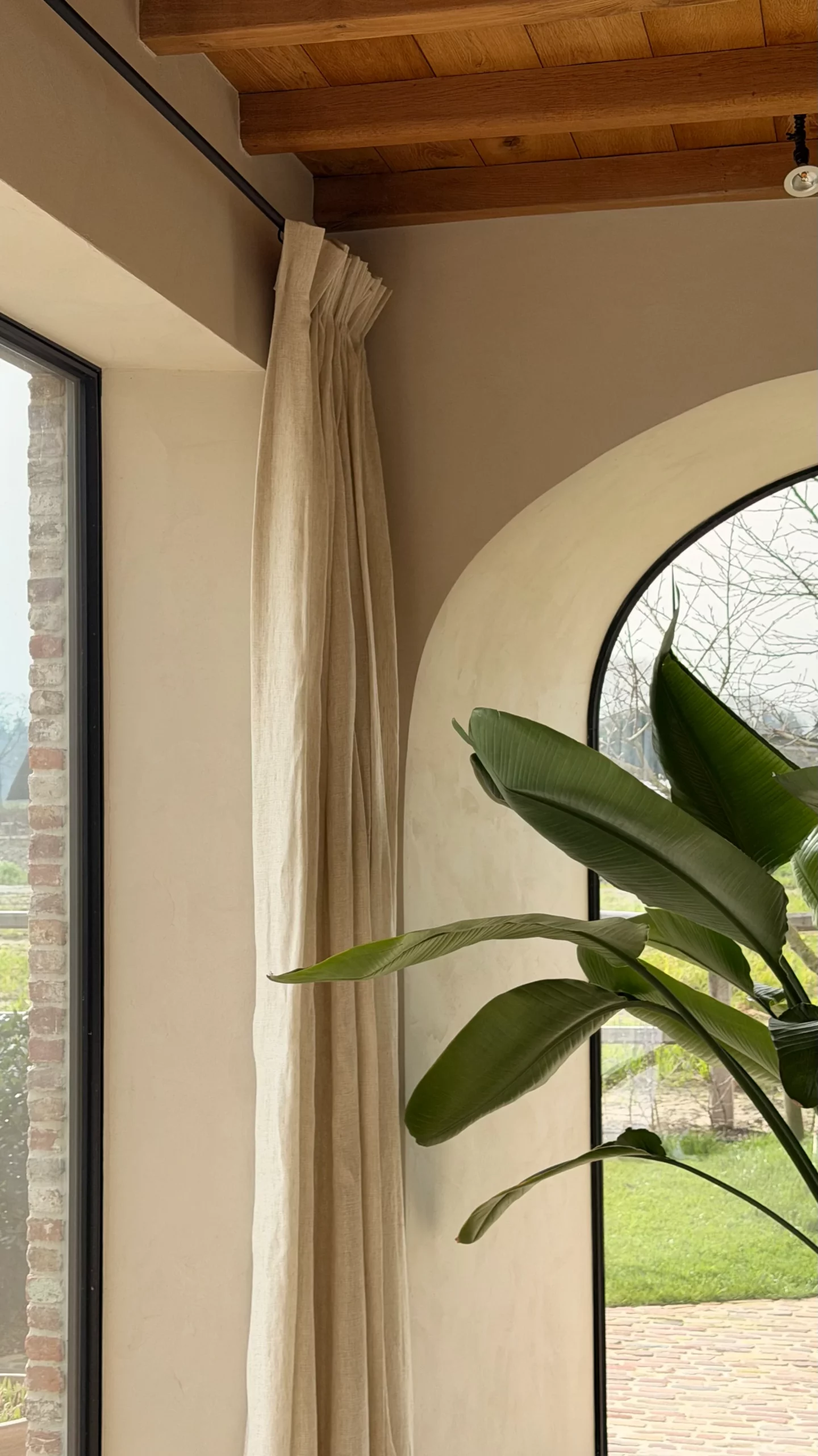 Beige linnen gordijn in een hoek naast een rond boogvenster in een mediterraan interieur met houten balkenplafond