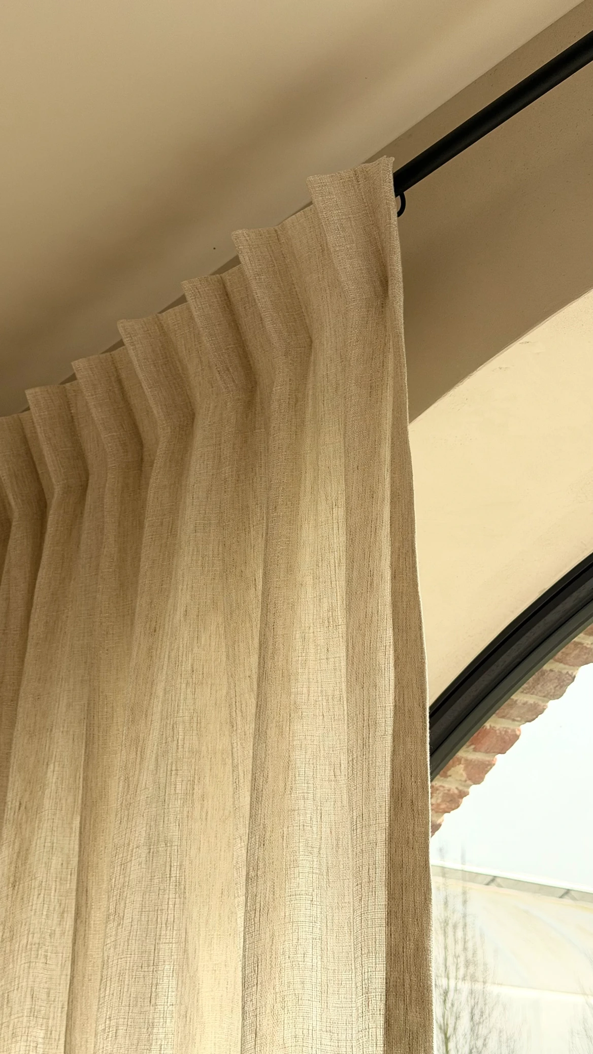 Close-up van een beige linnen gordijn met golfplooi op een zwarte gordijnrail bij een rond boogvenster