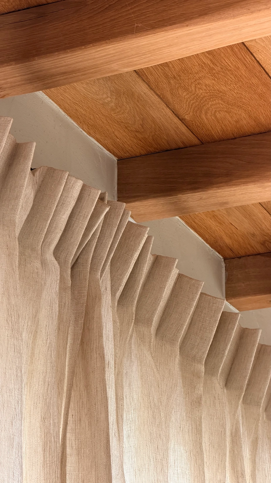 Beige linnen gordijnen met golfplooi tegen een houten balkenplafond, gefotografeerd van onderaf