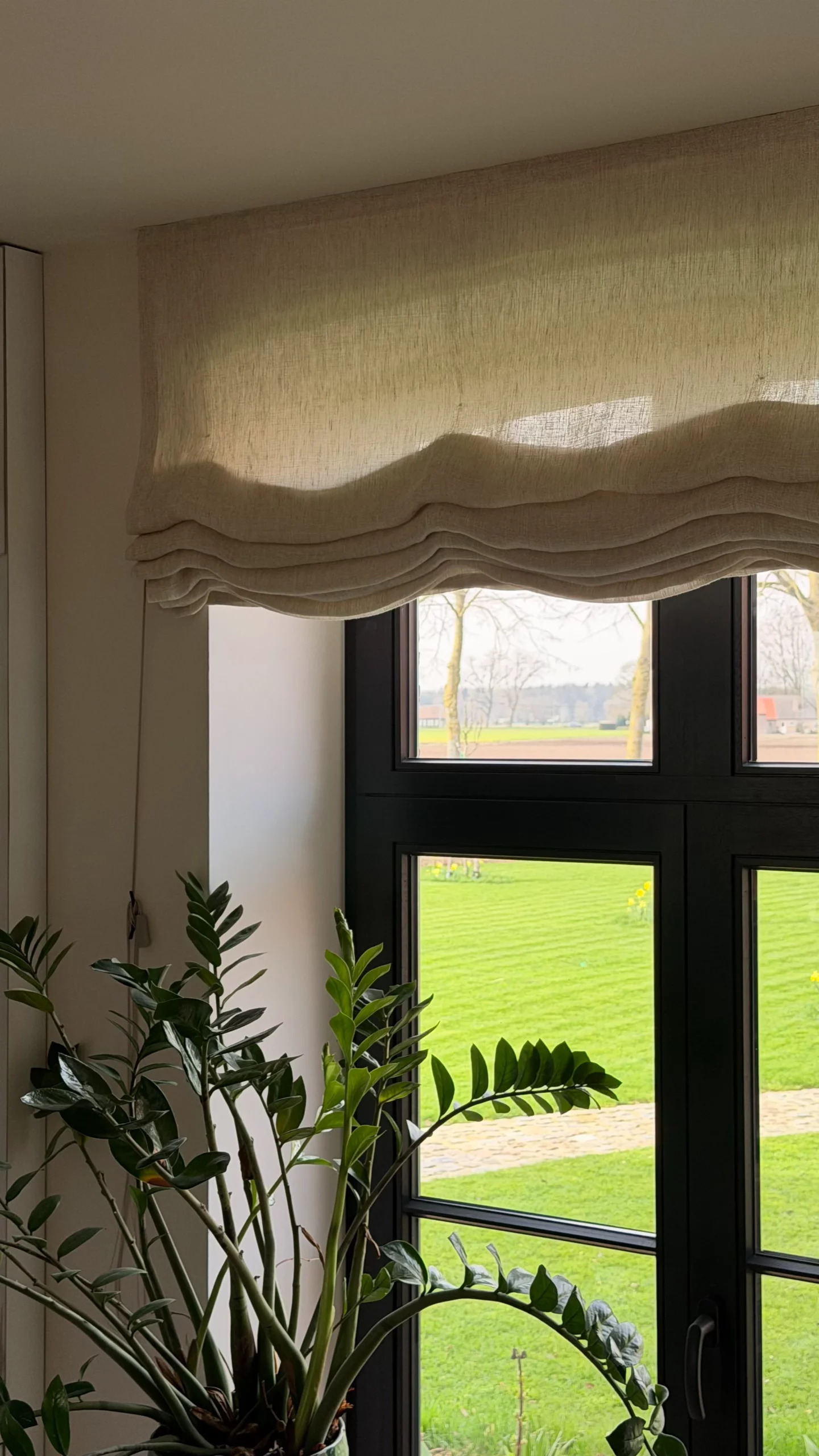Beige linnen vouwgordijn met zachte plooien boven een zwart raamkozijn met uitzicht op weilanden
