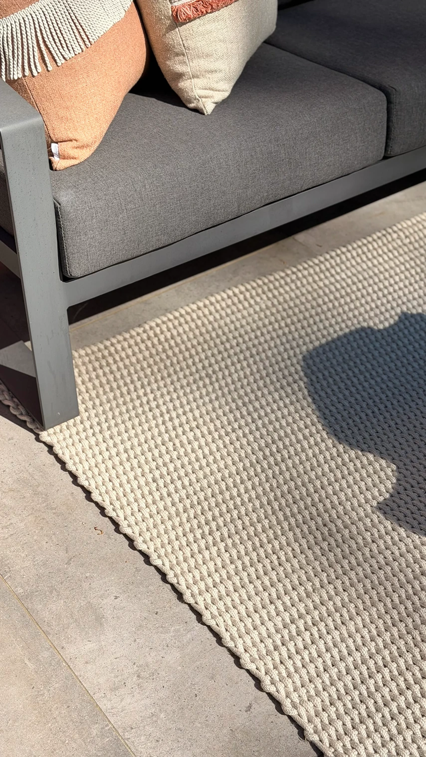 Close-up van een geweven beige buitenkleed onder een antracietgrijze buitenbank met terracotta sierkussens