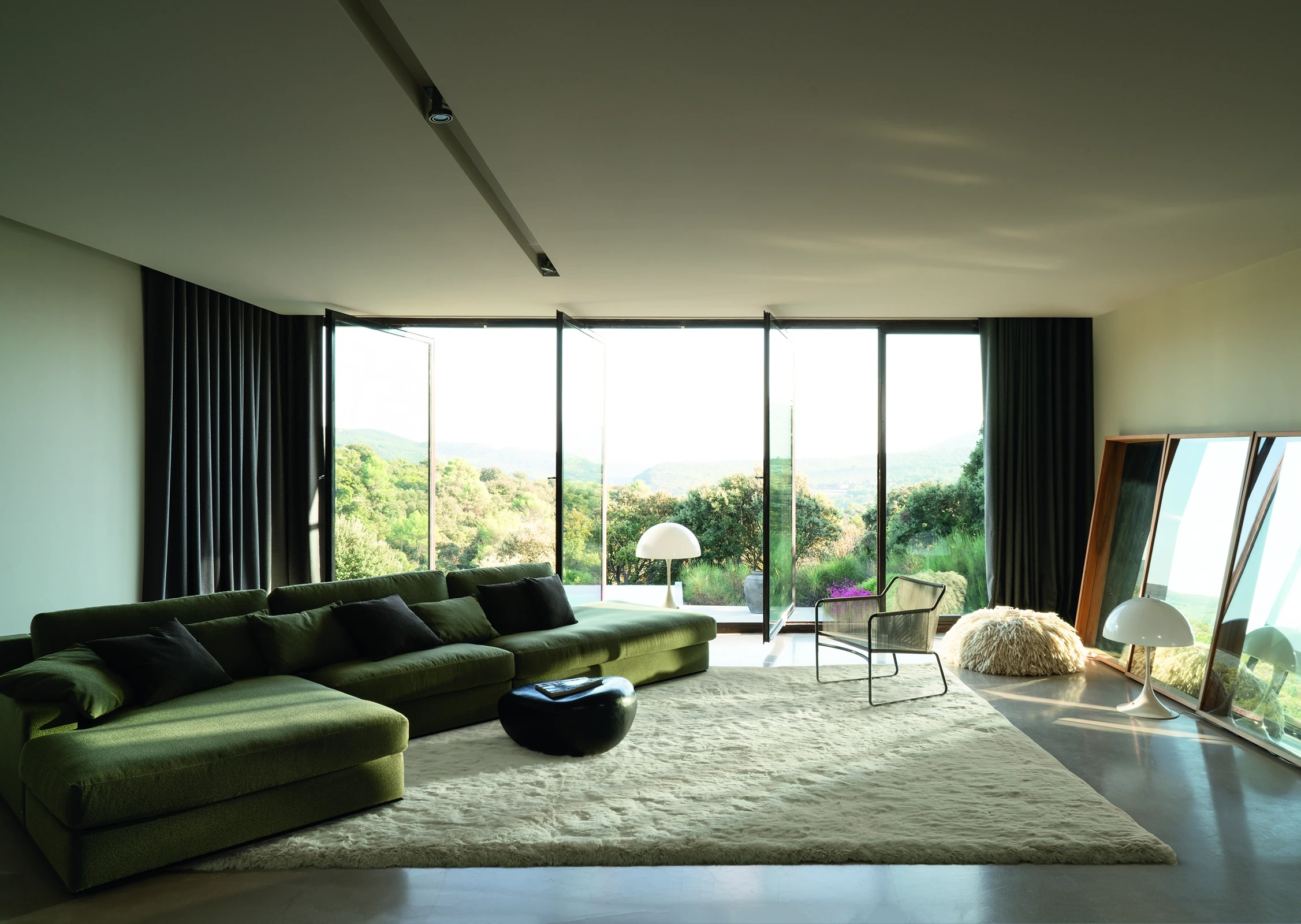 Donkere gordijnen bij een grote vouwwand met panoramisch heuvellandschap, in een woonkamer met olijfgroene loungebank