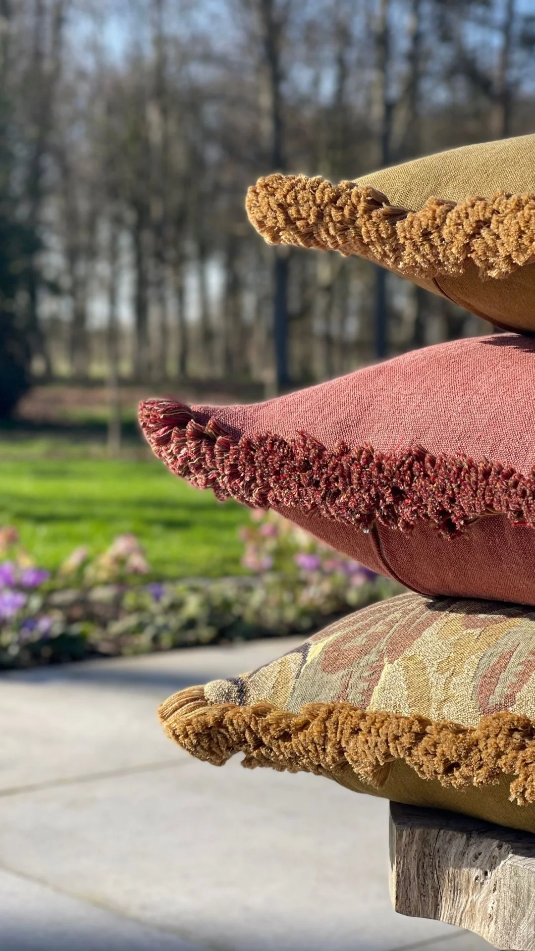 Drie gestapelde sierkussens in oker en roestrood met gedraaide franjerand, buiten bij een bloementuin