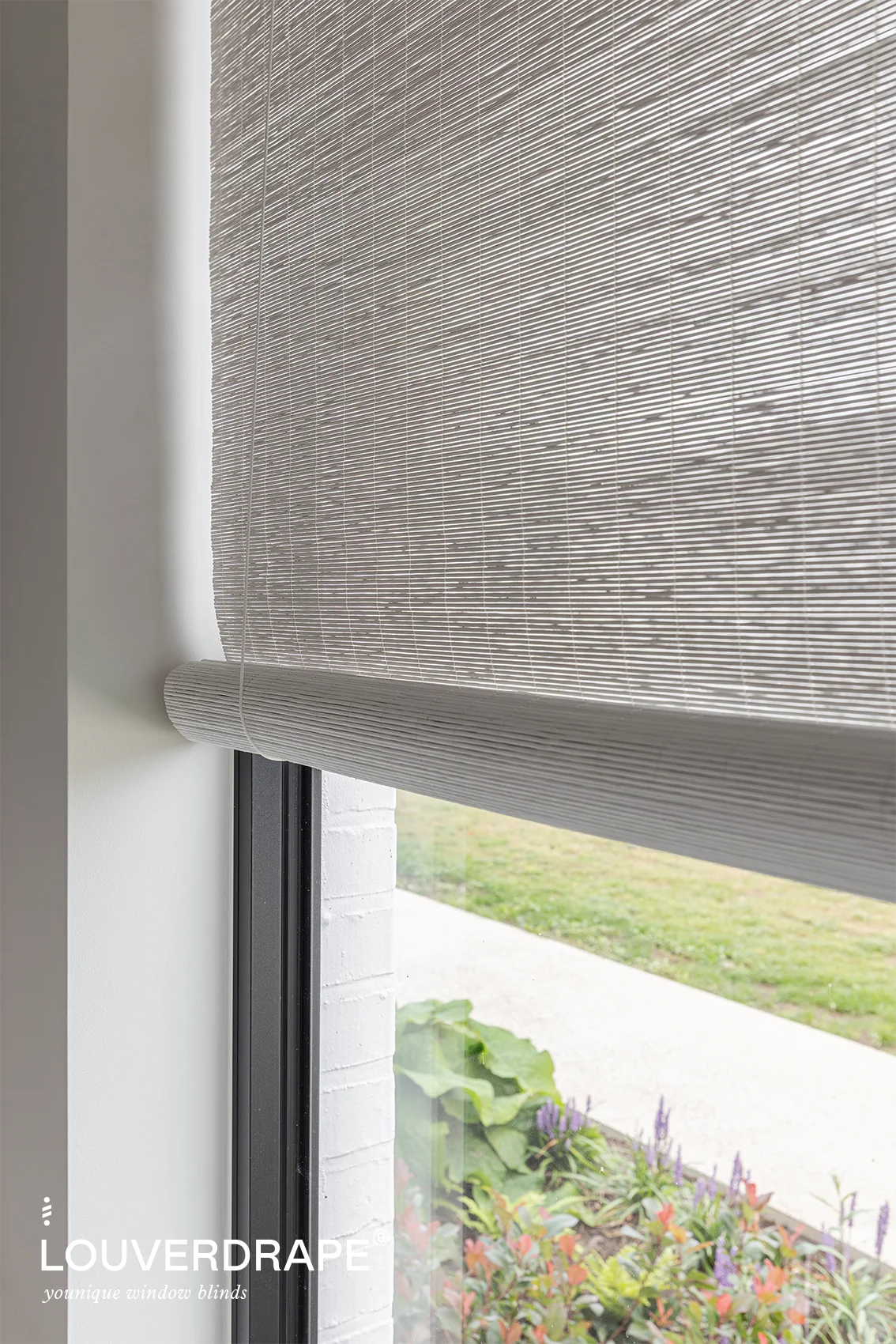 Close-up van een geweven houten rolgordijn van Louverdrape met zicht op een tuinpad met bloemen