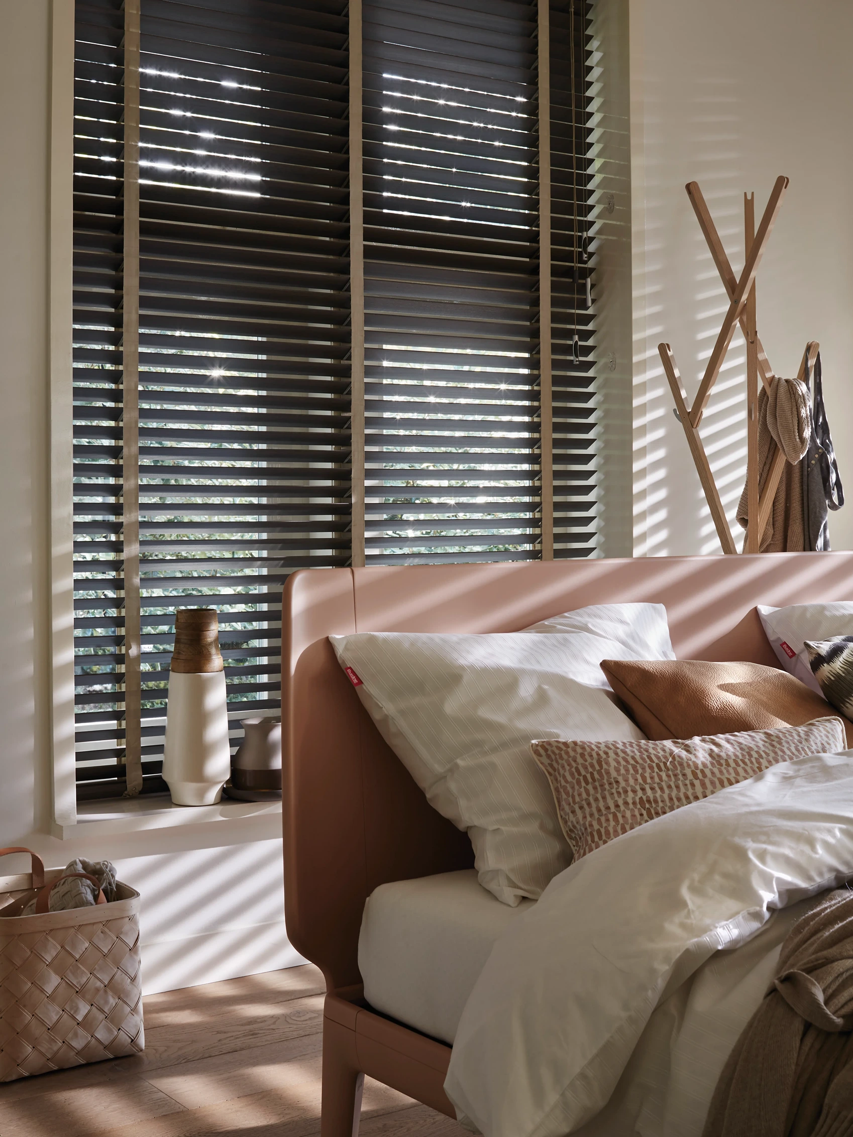 Houten jaloezieën met doorvallend zonlicht voor het raam van een slaapkamer met terracotta bed en beddengoed in aardetinten