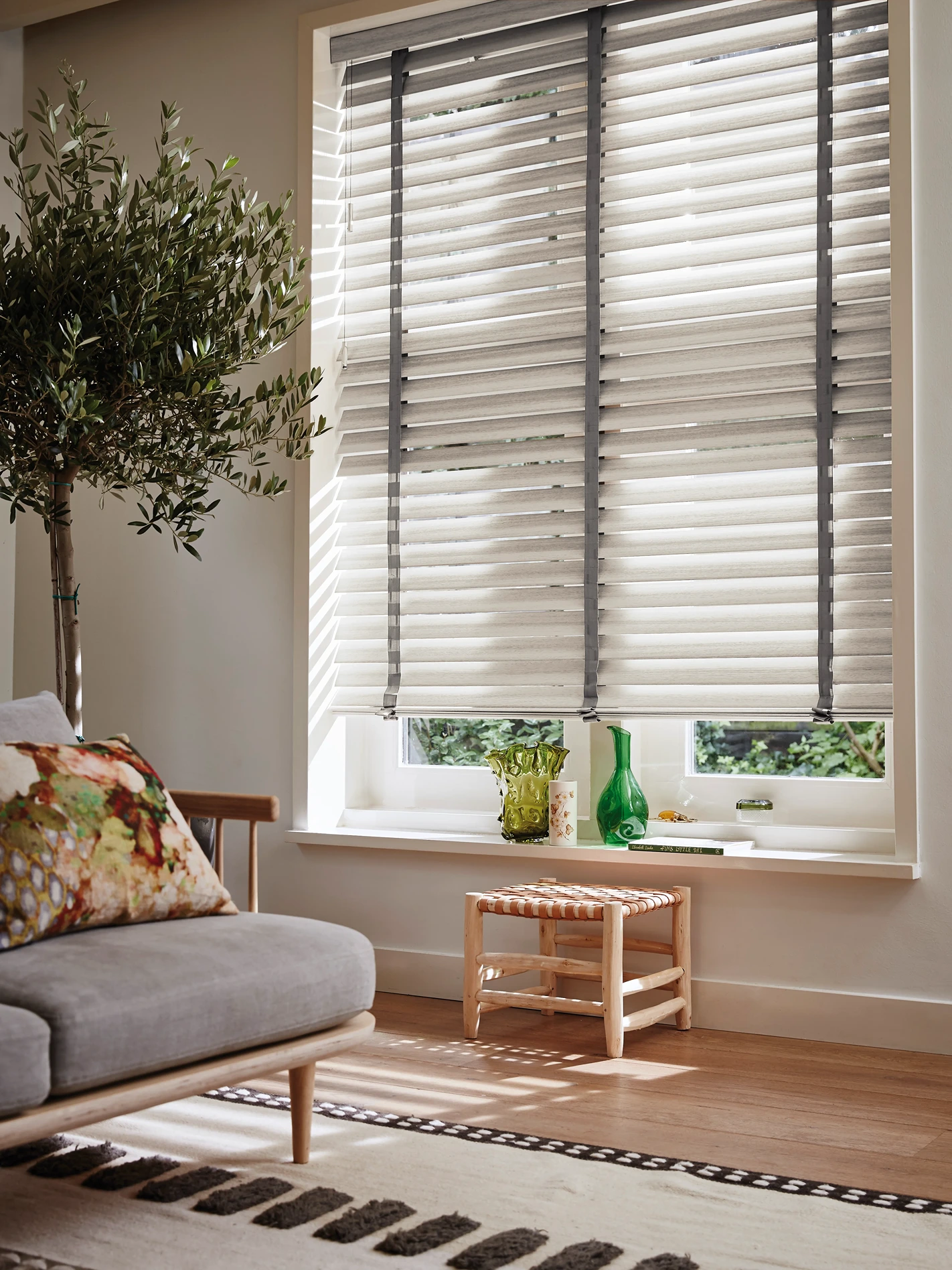 Houtlook jaloezieën met grijze bandjes voor een raam in een gezellige woonkamer met olijfboompje en grijze bank