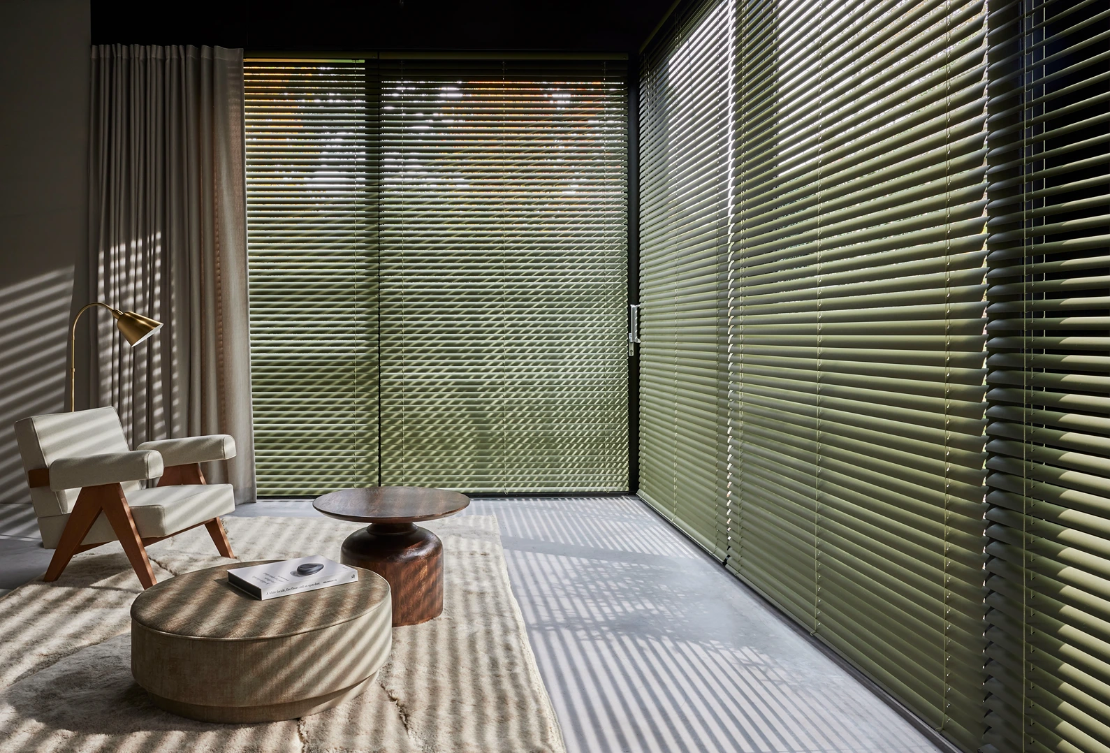 Olijfgroene aluminium jaloezieën over een glazen hoekwand met schaduwpatronen op de vloer, met design fauteuil en poef