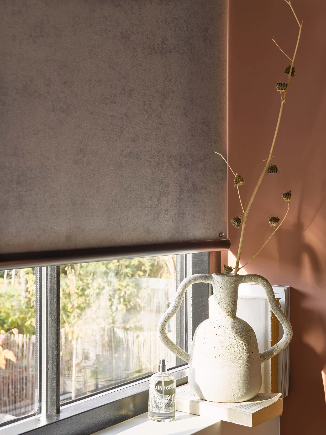 Taupe velours rolgordijn boven een vensterbank met keramische vaas en droogbloemen tegen een terracottakleurige muur