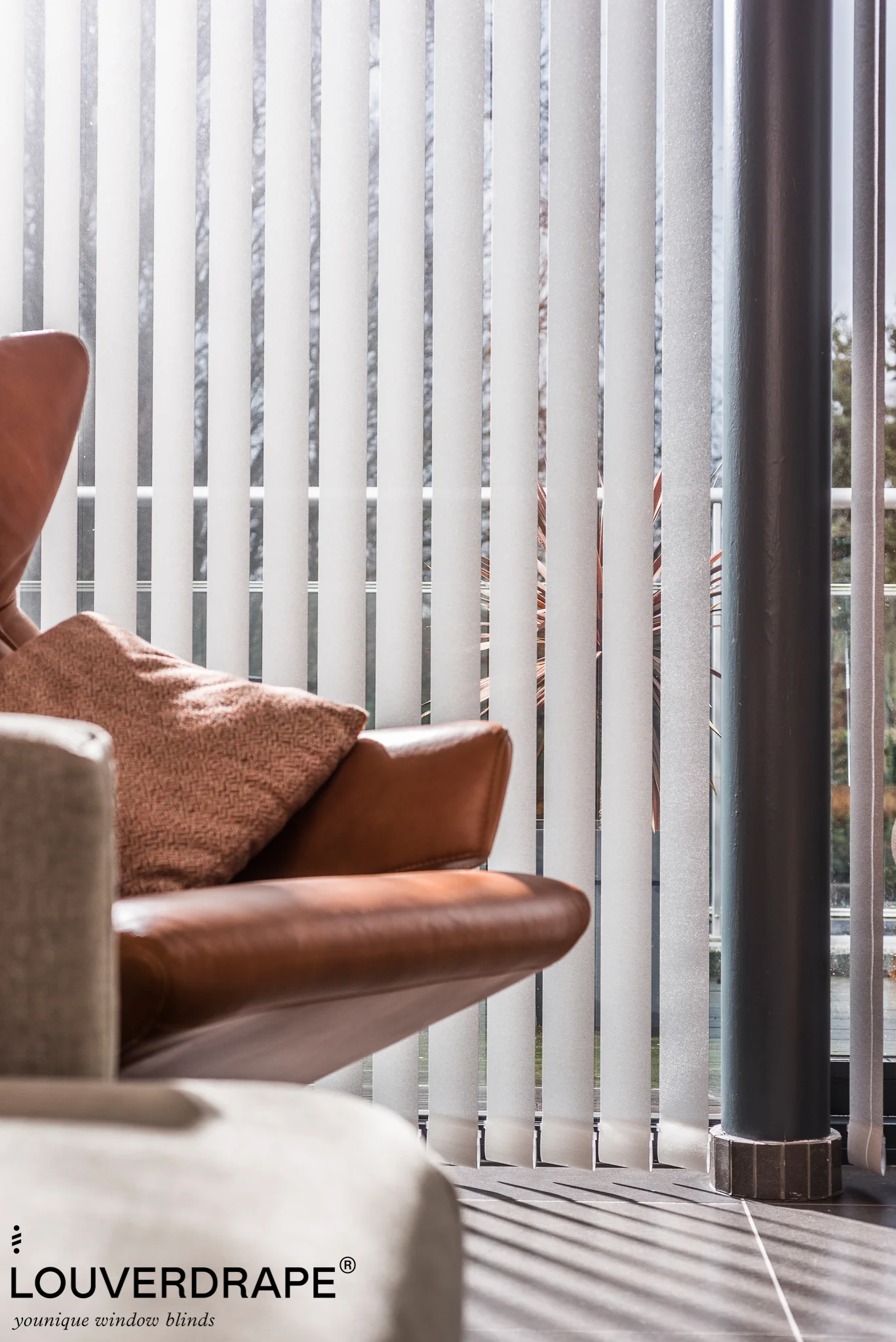Close-up van lichtgrijze verticale lamellen van Louverdrape voor een raam, met een bruine lederen fauteuil en terracotta kussen op de voorgrond