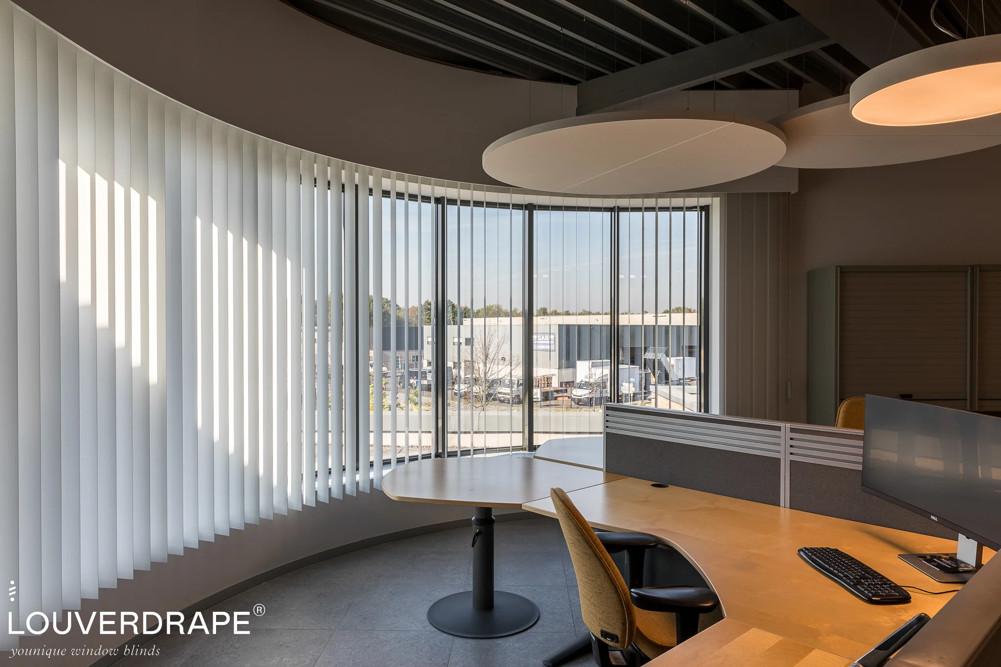 Witte verticale PVC-lamellen van Louverdrape voor een gebogen kantoorgevel met bureaus en zicht op een bedrijventerrein