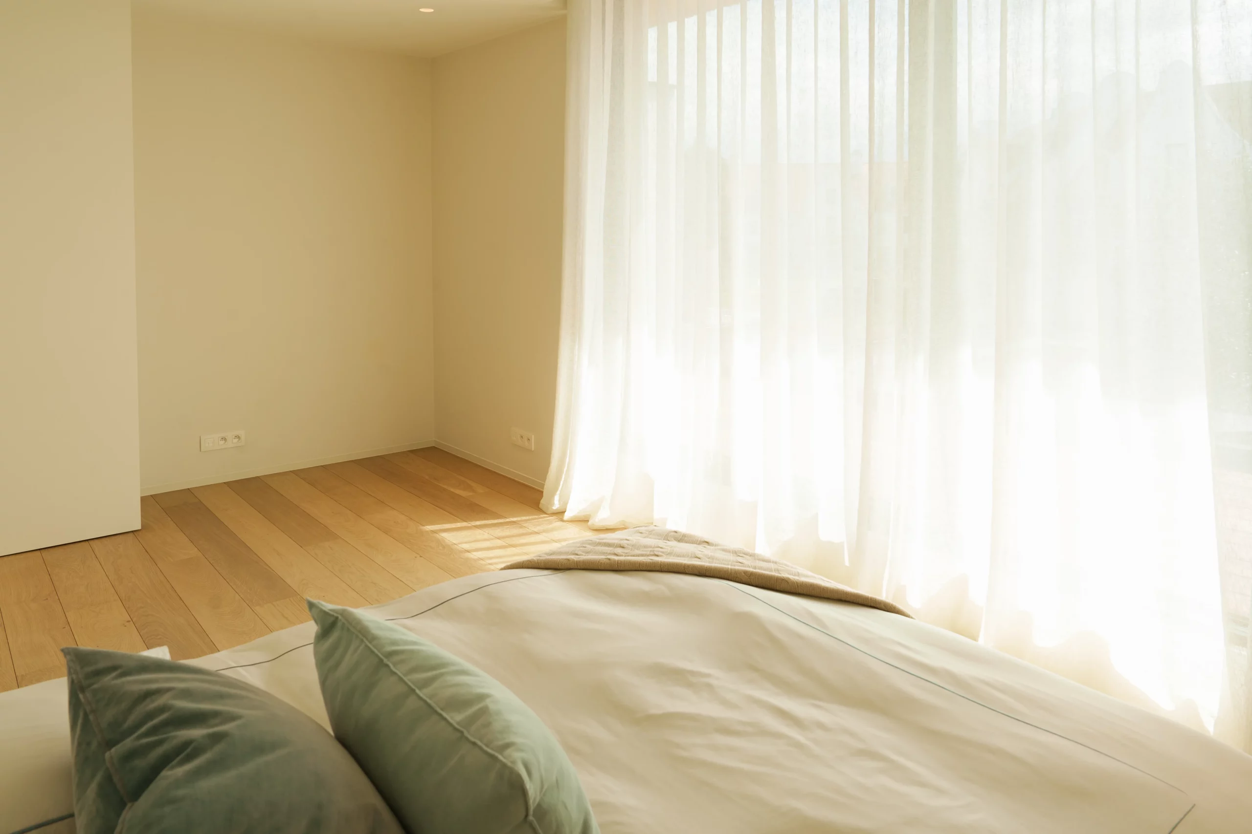 Wit vitragegordijn met golfplooi in een zonovergoten slaapkamer met wit dekbed en salie-groene kussens