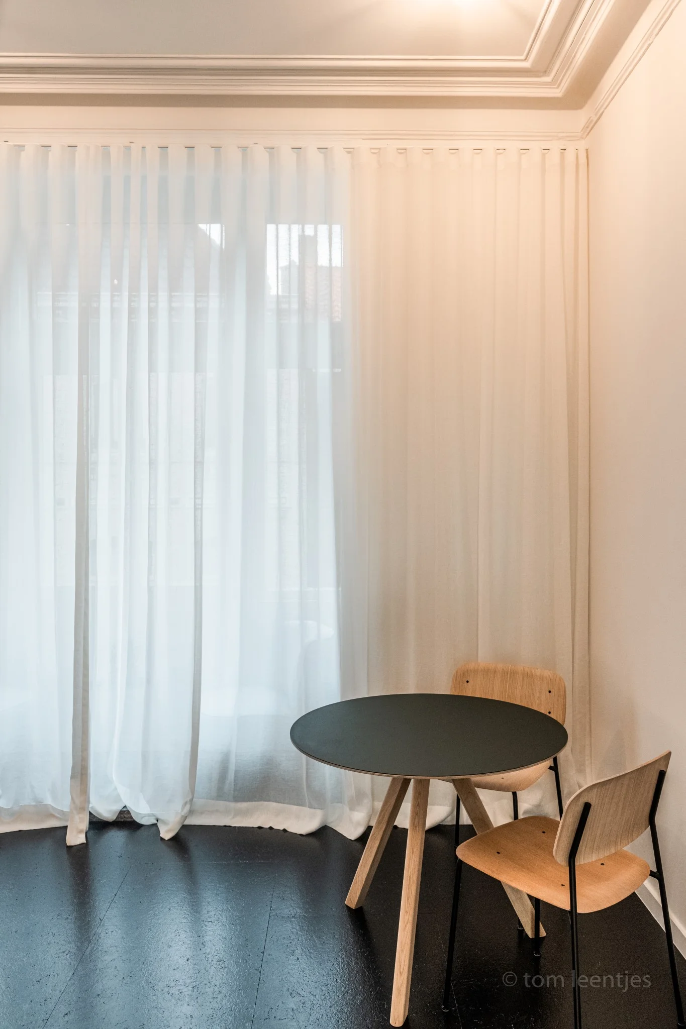 Witte golfplooigordijnen tot op de vloer in een eethoek met ronde tafel, houten stoelen en donkere houten vloer
