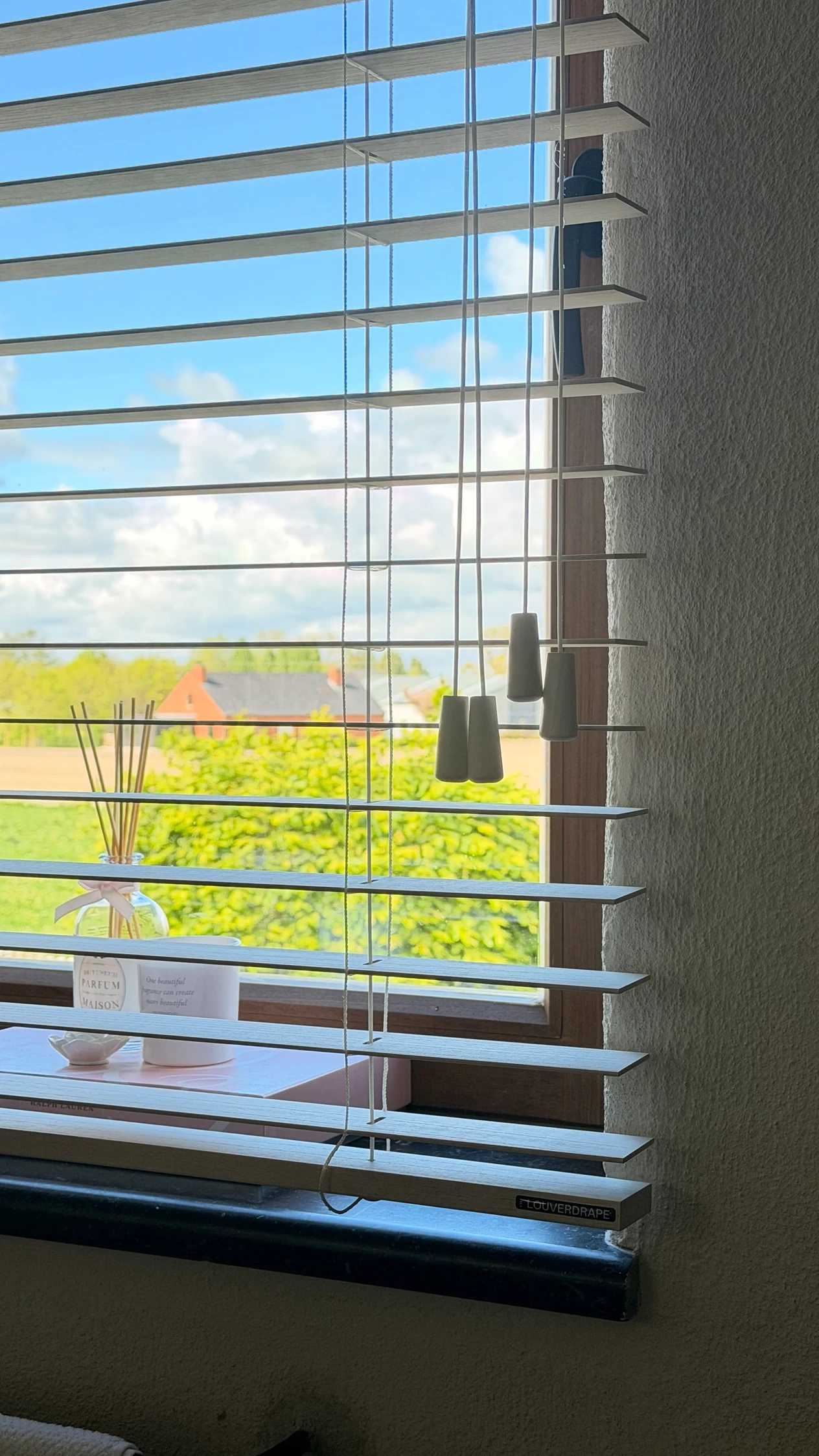 Witte houten jaloezieën van Louverdrape voor een raam met zonnig landelijk uitzicht en vensterbank met geurstokjes